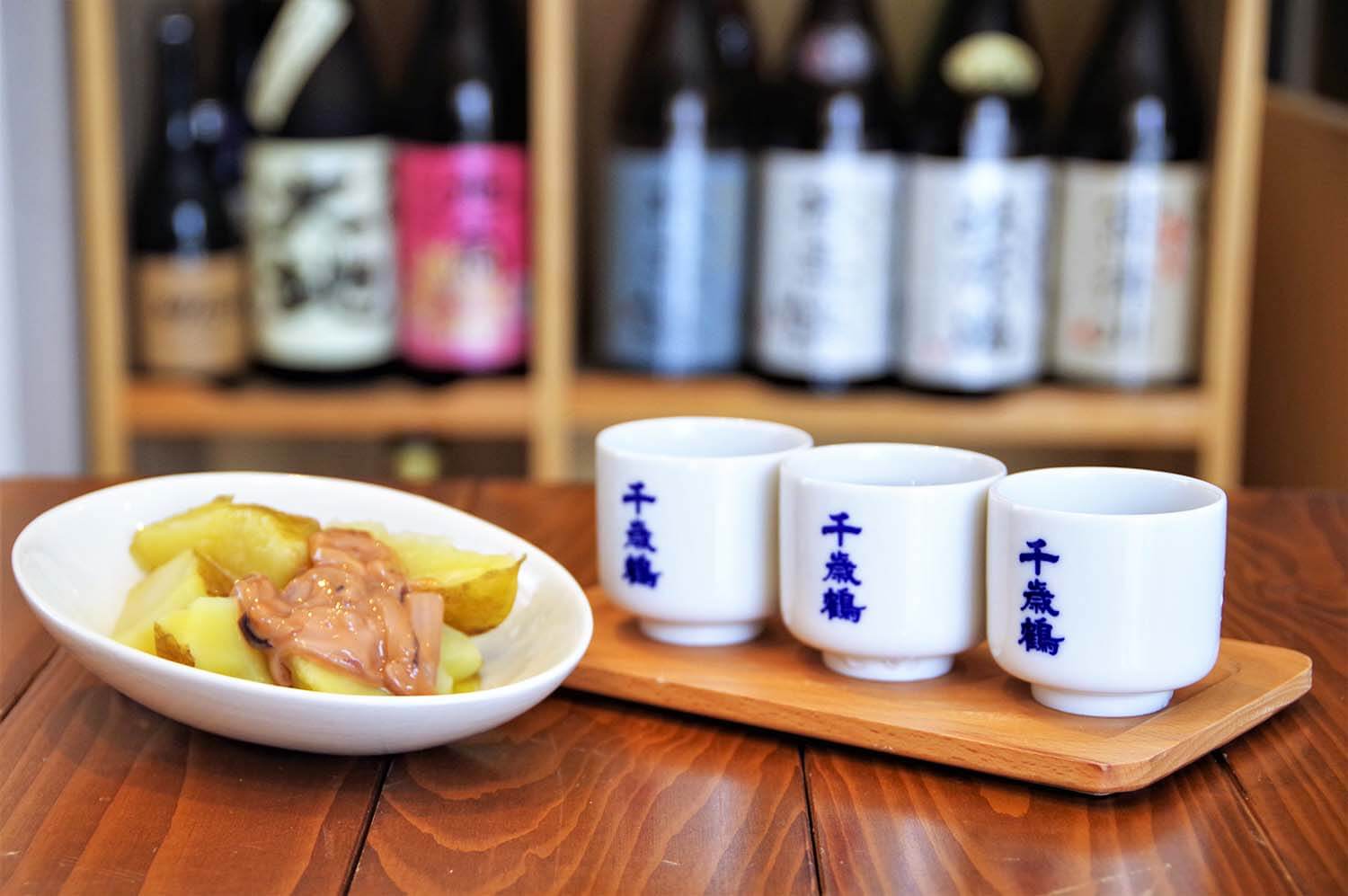 「日本酒3種飲み比べ」600円と「塩辛じゃがバター」550円。角打ちの日本酒は1合300円～